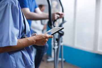Close-up of nurse using a tablet at clinic, reading a patient report while wearing blue scrubs and stethoscope. Caucasian female physician holding digital device for medical research.