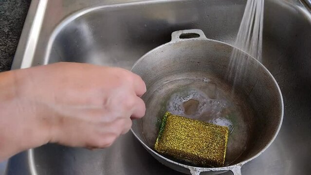 Unas manos de mujer lavando  un caldero  con una esponja amarilla con jab&oacute;n l&iacute;quido y  agua en un fregadero