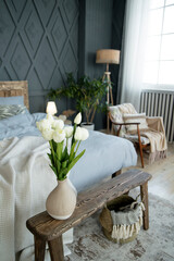 Wooden bench with a vase of white tulips as the main focus. Surrounding elements include a cozy bed with blue linens, sheer curtains, and a wicker basket on a patterned rug