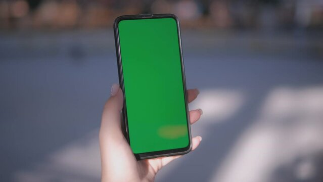 Hand holding smartphone with green screen, perfect for mockups or chroma key effects, blurred outdoor setting.