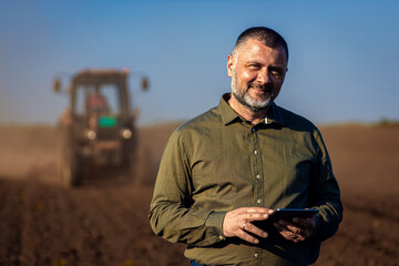 Portrait of satisfied mature farmer standing in field with tablet supervises the cultivation of...