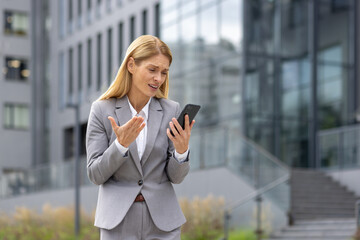 Fototapeta premium Upset young business woman in a gray suit standing on the street outside an office building and looking frustrated at the mobile phone screen.