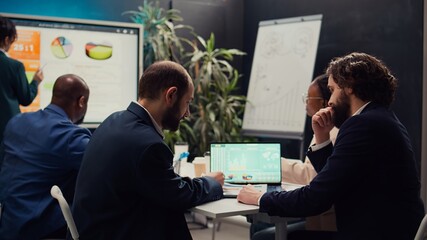 Business partners examining sales infographics in a boardroom, reviewing market trends and competitive analysis. Specialists share ideas for new products, ensuring company success. Camera B.