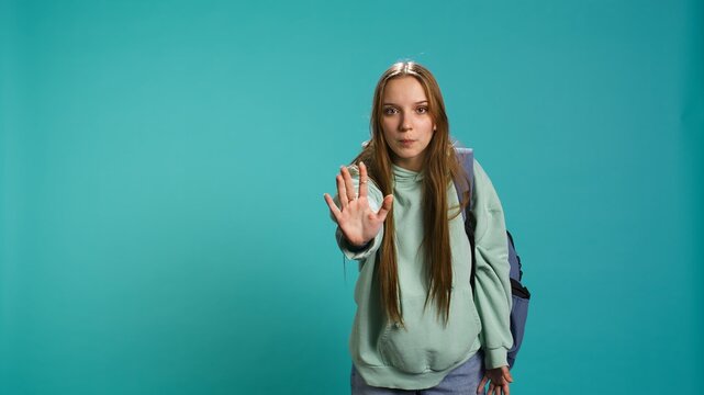 Assertive young woman doing stop hand gesture and talking. Annoyed girl doing firm halt sign gesturing, wishing to end concept, isolated over blue studio background, camera B