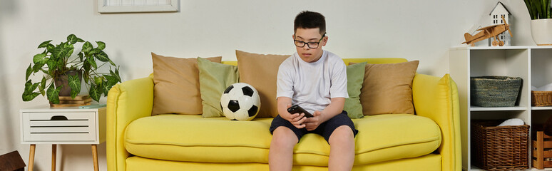 A young boy sits on a yellow couch, holding a phone, with a soccer ball nearby.