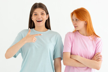 Portrait of two cheerful young women standing together, one girl point at the other, the other one is unhappy isolated over white background