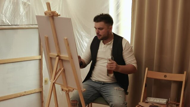 A young artist sits at home on a stool in front of a canvas and works with inspiration, paints a picture, and drinks alcohol from a bottle, happy to do what he really likes,