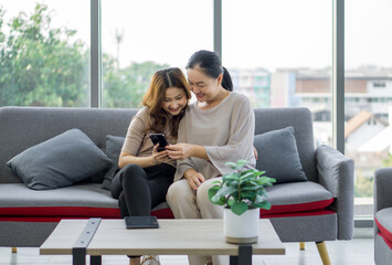 Obraz premium Two asian women sitting on a couch, smiling and looking at a mobile phone together.