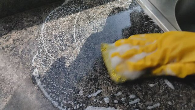 Primer plano de una mano de mujer con un guante amarillo y una esponja con jab&oacute;n limpiando la cocina de su casa.