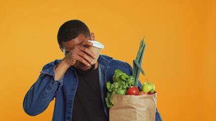Young adult enjoying a cup of coffee during his grocery shopping spree, carrying a paper bag full of organic produce in studio. Man serving caffeine refreshment, zero waste. Camera A.