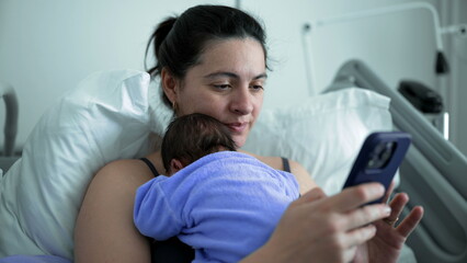 New mother in hospital bed holding smartphone and newborn baby, multitasking between technology and...