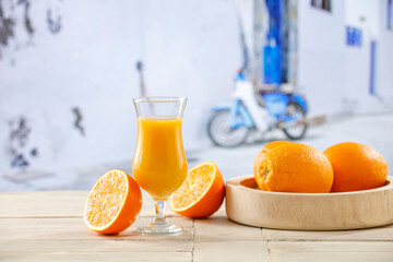 Glass of fresh orang juice with oranges cut in halves on wooden plate on wooden table and blurred city background in distance.