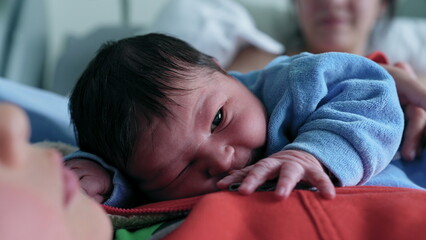 Newborn baby resting on older sibling's chest, looking alert with mother in background, capturing a precious family bonding moment in a warm and loving environment