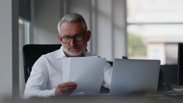 Serious adult businessman checking corporate paperwork correspondence sitting at home office desk. Male entrepreneur reading documents, analyzing financial papers, preparing audit report at workplace.