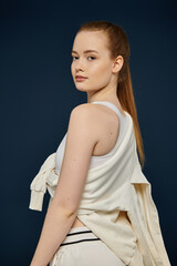 Young woman in white tank top and draped sweater poses confidently on deep blue backdrop.