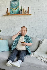 A man with inclusivity sits on a couch, talking on the phone and holding a credit card.