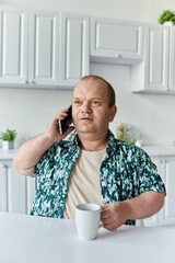 A man with inclusivity in a green patterned shirt talks on his phone while holding a white mug.