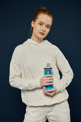 Woman in white hoodie holds blue water bottle against blue backdrop.