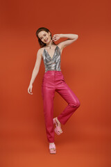 Confident woman in silver chainmail top and pink leather pants on orange background.