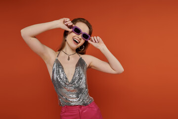 A young woman in a silver top and pink pants poses with sunglasses against an orange background.