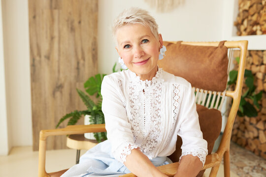 Stylish mature successful lady entrepreneur of 60s spending her vacation in cozy country house of boho style, looking at camera sitting in stylish chair in laced blouse and earring of exclusive design