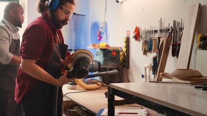 Carpenter at work consulting blueprint, using orbital sander on lumber, wearing safety glasses. Artisan with protecting equipment uses angle grinder on wood after checking schematics, camera B