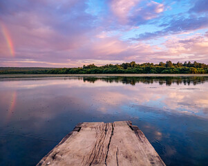 Beautiful pink sunset with rainbow reflection over calm lake at countryside evening. Tranquil lake with a wooden dock in the foreground.