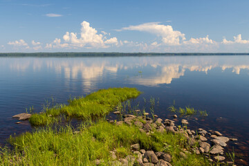 Beautiful summer landscape. View from the shore to the lake. Windless weather, calm. Clouds are reflected in the water. Rocky shore covered with grass. Lake Shotozero, Republic of Karelia, Russia.