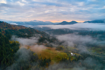 Mountains in clouds at sunrise in summer. Aerial view of mountain peak with green trees in fog. Top view from drone of mountain valley in low clouds