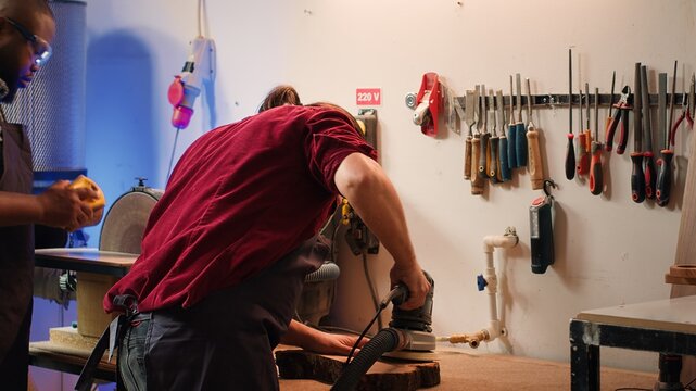 Carpenter collaborating with apprentice, using orbital sander with fine sandpaper to achieve refined finish. Manufacturer and BIPOC colleague using angle grinder on wood block, camera A