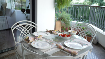 Elegantly Set Dining Table on Balcony with Fresh Tomato Salad, Surrounded by Green Plants and Modern Home Decor