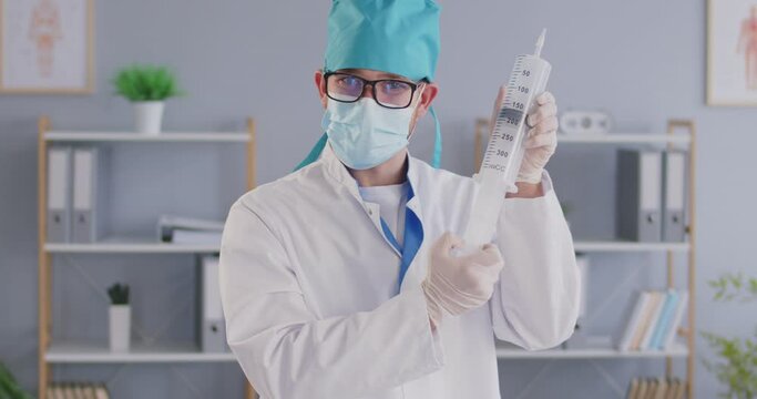 Video portrait funny man professional nurse or doctor in white uniform coat, green scrubs hat, blue facial mask, glasses and surgical latex gloves holding big enormous syringe ready for vaccine shot
