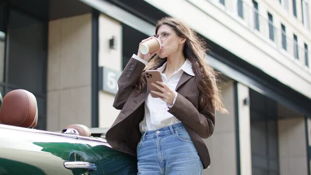 A businesswoman pauses outdoors, multitasking with her cellphone and coffee, connecting with the camera in a moment of brief respite