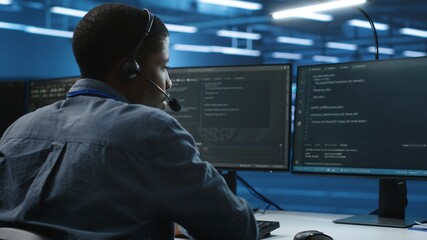 IT support worker in data center talking in headset mic with coworkers, coding on computer. Man...
