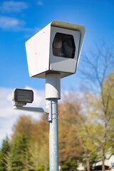 Traffic surveillance camera on a metal pole against a clear blue sky and blurred trees in the background.