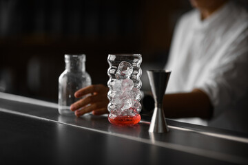 Focus on a shaped glass filled with ice cubes, next to which on the bar stand a jigger and a jar held