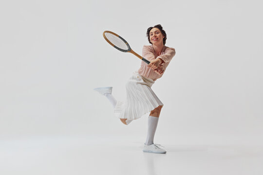 Dynamic image of happy, smiling, beautiful young woman in skirt and blouse cheerfully playing tennis isolated on white background. Concept of sport, retro and vintage, active lifestyle, hobby