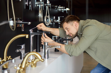 Man in a plumbing store, comparing different taps and faucets in preparation for a bathroom renovation at home. He evaluates each option in shop to find the perfect fit for plumbing needs and design.