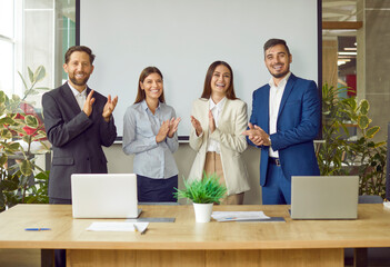 Happy business team clap hands after successful presentation at work meeting. Portrait of smiling applauding people standing by table in modern office boardroom with green plants and projection screen