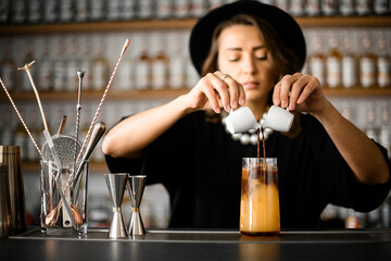 Cute female bartender pouring cinnamon drink from two white cups into tall glass with juice