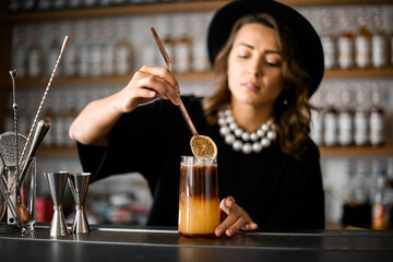 Cute female bartender drops an orange slice into a juicy orange-brown cocktail with ice in a tall glass