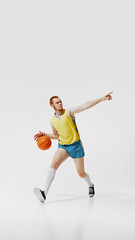 Full-length image of young man in yellow jersey and blue shorts, basketball player in motion, training isolated on white background. Concept of sport, retro and vintage, active lifestyle, hobby