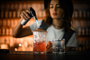 Female bartender holds a lit gas burner in her hands, pointing it at the ceramic lid