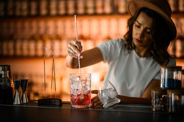 Female bartender stirs pink cocktail with ice in mixing glass with long bar spoon