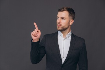 Caucasian millennial businessman isolated on gray studio background posing, satisfied successful man in formal suit pointing something with finger at copy space