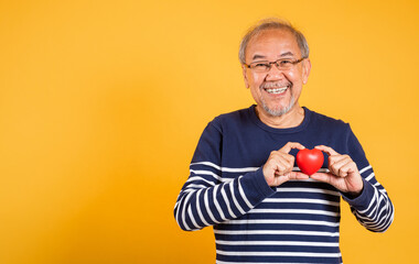 Portrait Asian smiling old man holding a red heart in his hand studio shot isolated yellow background, Excited happy romantic senior man pensioner filling lover, Concept of happiness and warmth