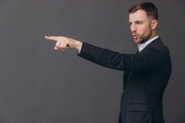 Caucasian millennial businessman isolated on gray studio background posing, nervous angry man in formal suit standing in profile pointing finger and saying you copy space