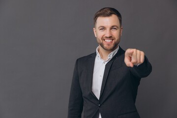 Caucasian millennial businessman isolated on gray studio background posing, smiling successful man in formal suit pointing finger and saying you on copy space