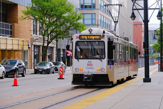 An RTD light rail train travels through a Denver city street lined with buildings, trees, and parked cars, with pedestrians walking along the sidewalk.	
