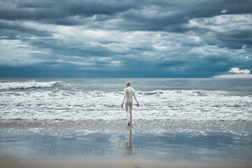 Hairless girl with alopecia in white futuristic suit goes to cold restless sea on sandy beach,...